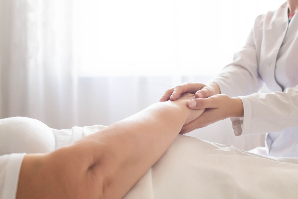 Doctor holds the hand of an elderly woman patient on a background of a window. Concept of hygienic patient care in a hospital, geriatrics, copy space