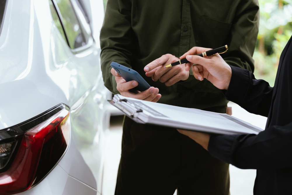 Insurance agent reviews car accident claim. Agent holds clipboard as customer signs form. Both discuss financial arrangements for damaged vehicle to ensure successful service.